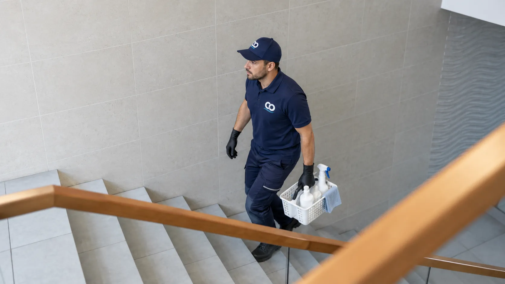 Mann in dunkelblauer Arbeitskleidung trägt einen Korb mit Reinigungsmitteln eine helle Steintreppe hinauf